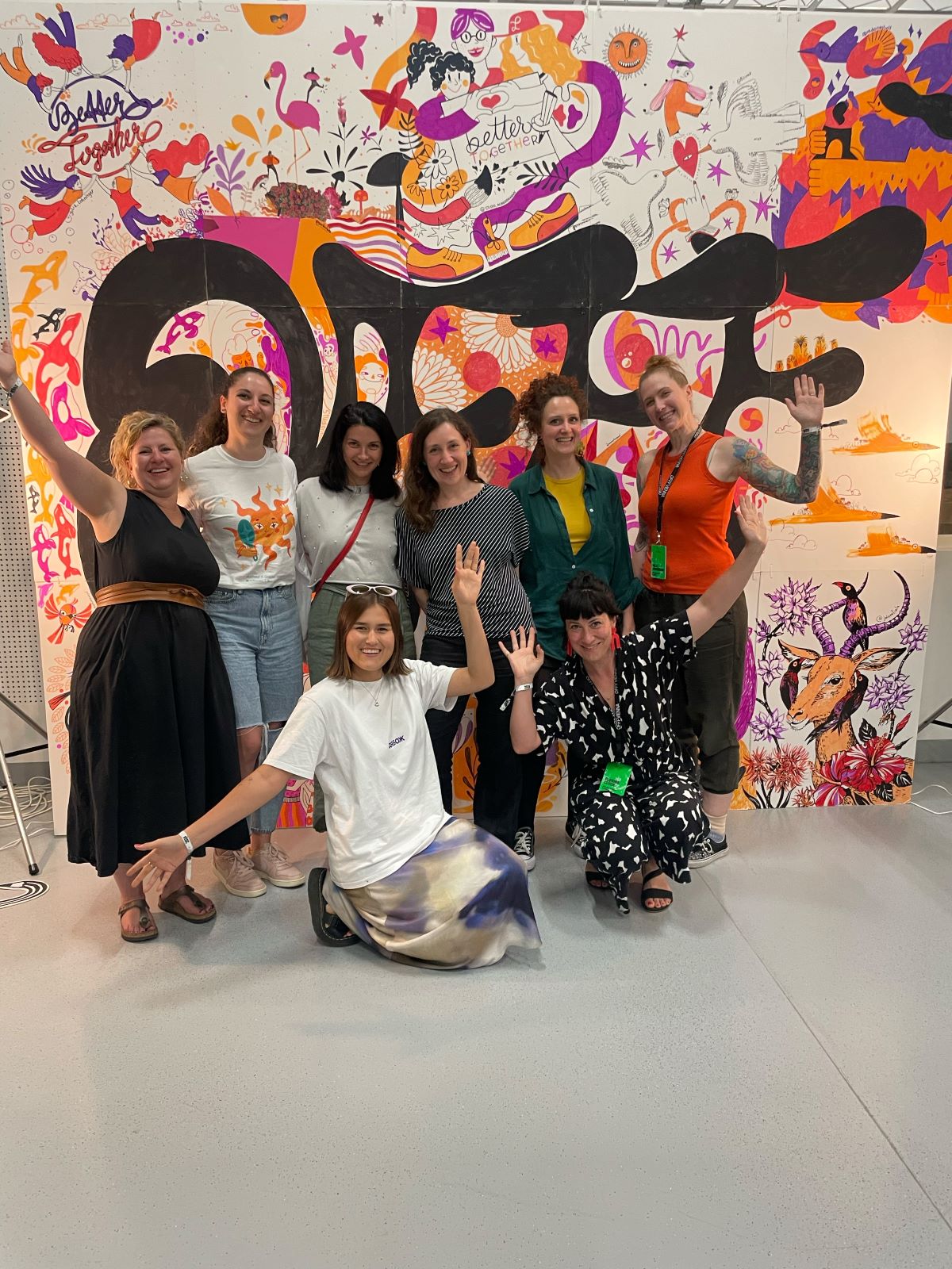 Acht Frauen lächeln und posieren mit erhobenen Armen vor einem farbenfrohen, illustrierten Hintergrund der Illustration Ladies bei der Veranstaltung von Stift und Haltung in Wien. Die Atmosphäre ist fröhlich und lebendig, Kreativität und Stolz werden gefeiert.