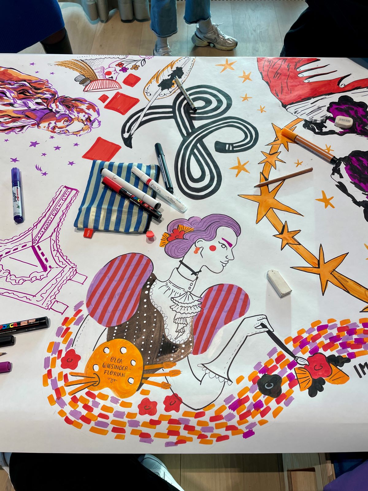 Ein großes Blatt Papier zeigt farbenfrohe Illustrationszeichnungen, darunter eine Frau in historischem Kleid, wirbelnde Muster, Sterne und Blumen. Um das Kunstwerk herum sind Hände und Marker zu sehen, was auf eine gemeinsame künstlerische Tätigkeit von Frauen in Wien hindeutet.