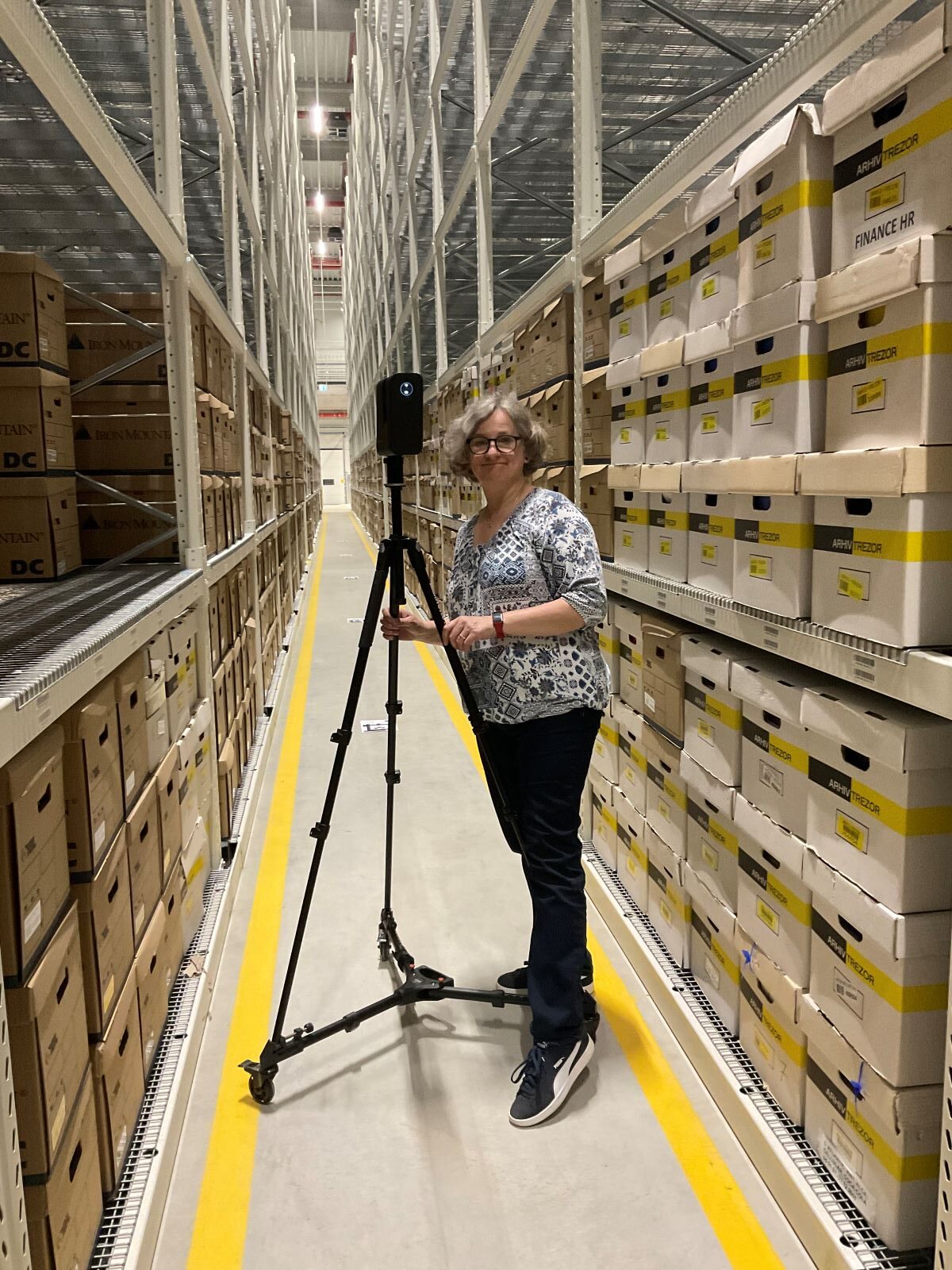 Eine Person steht in einem Lagergang zwischen Regalen mit Archivkartons und hält eine auf einem Stativ montierte Kamera oder einen Scanner. Der Bereich ist gut beleuchtet und geordnet, die Kisten sind beschriftet und ordentlich gestapelt.