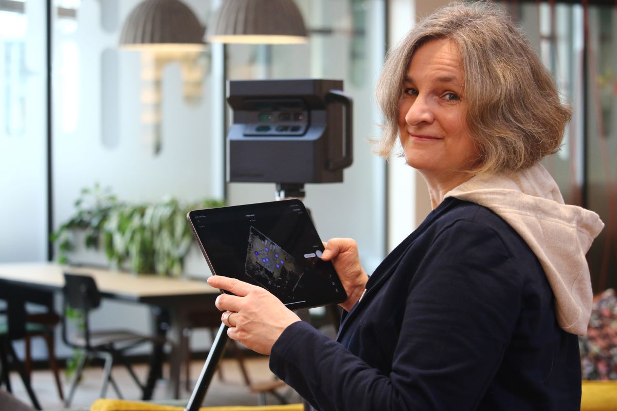 Eine Frau mit grauem Haar lächelt, während sie ein Tablet hält und neben einer 3D-Kamera in einem modernen Büro mit Pflanzen und Holzmöbeln im Hintergrund steht.