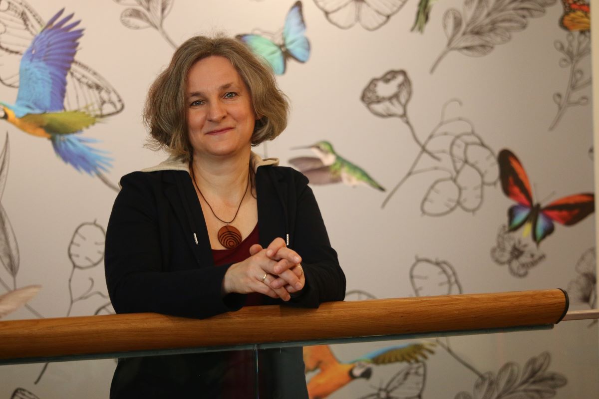 Eine Frau mit kurzem, gewelltem Haar steht hinter einem Holzgeländer und lächelt. Sie trägt eine dunkle Jacke und eine Halskette. Im Hintergrund ist eine mit bunten Schmetterlingen, Vögeln und Blumenmustern verzierte Wand zu sehen.