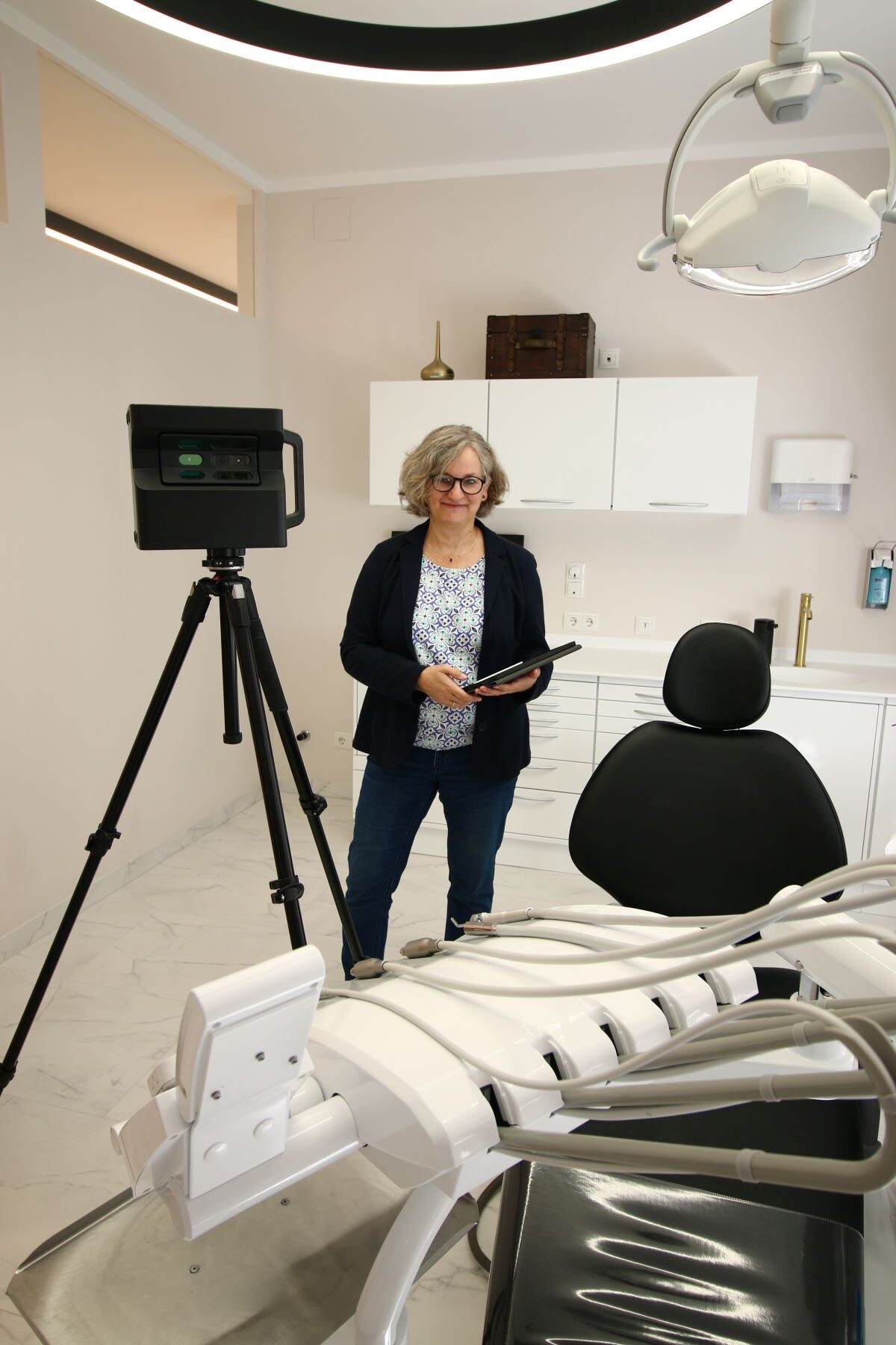 Eine Frau steht in einer modernen Zahnarztpraxis und hält ein Tablet neben einer Kamera auf einem Stativ. Im Vordergrund sind ein leerer Zahnarztstuhl und Geräte zu sehen, im Hintergrund stehen Schränke und Lampen.