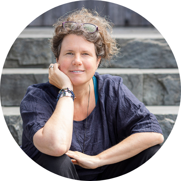 207 Eine Person mit lockigem Haar, einer Brille auf dem Kopf, einem dunklen, weiten Oberteil und Armbändern sitzt auf den Steinstufen im Freien, lächelt und stützt das Kinn auf die Hand.