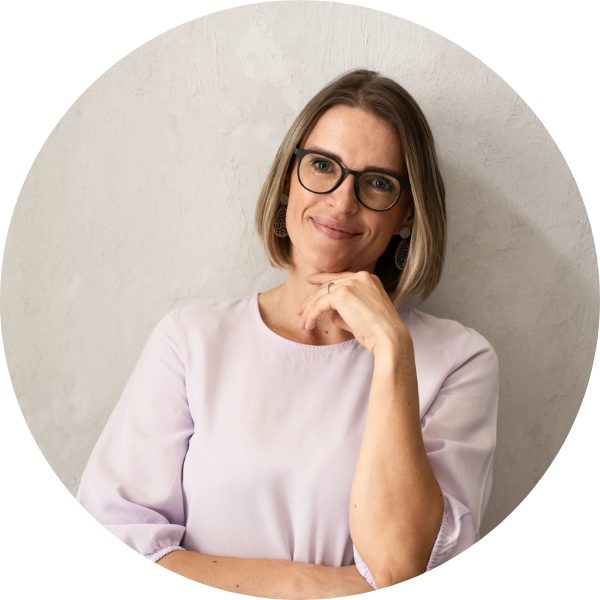 207 Eine Frau mit schulterlangem blondem Haar und Brille, die ein hellviolettes Oberteil trägt, lächelt sanft und stützt ihr Kinn auf die Hand, während sie vor einer hellen, strukturierten Wand steht.