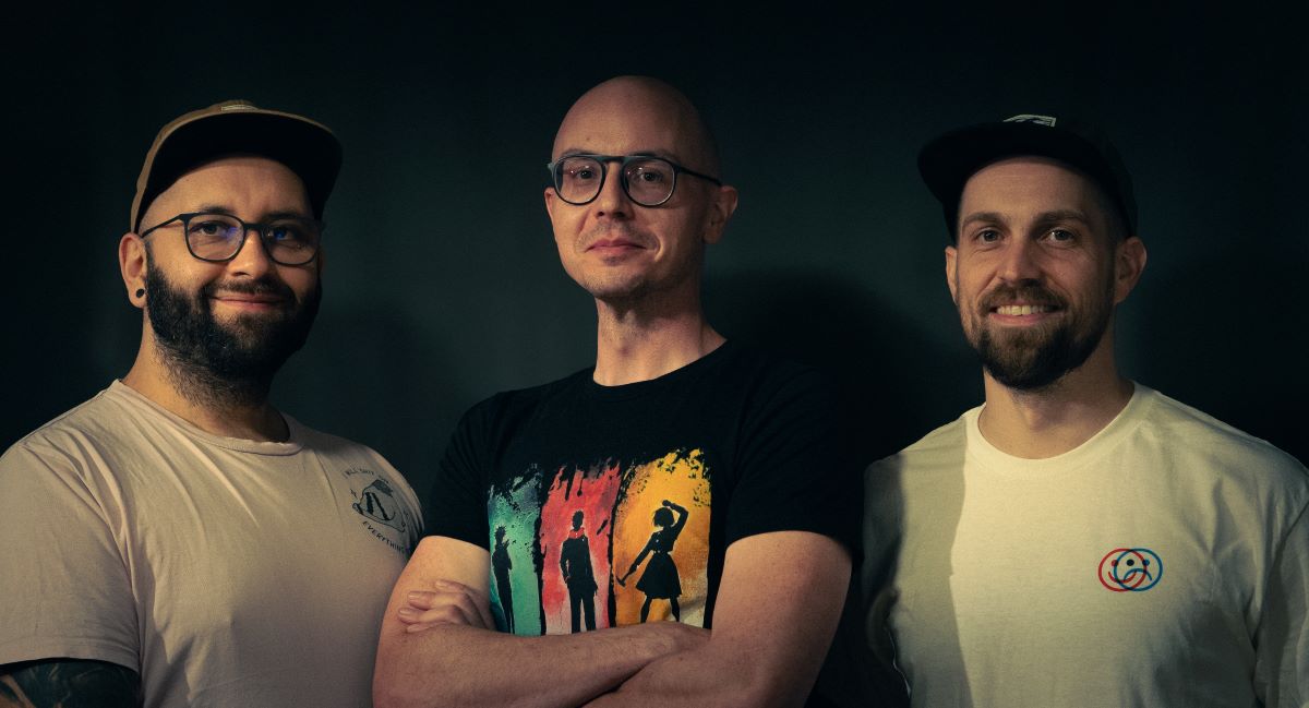 Three men stand together in front of a dark background and smile in the style of Hollywood quality on an SME budget. The man in the middle is bald, wears glasses and a black T-shirt; the others have beards, glasses, light-colored T-shirts and caps.