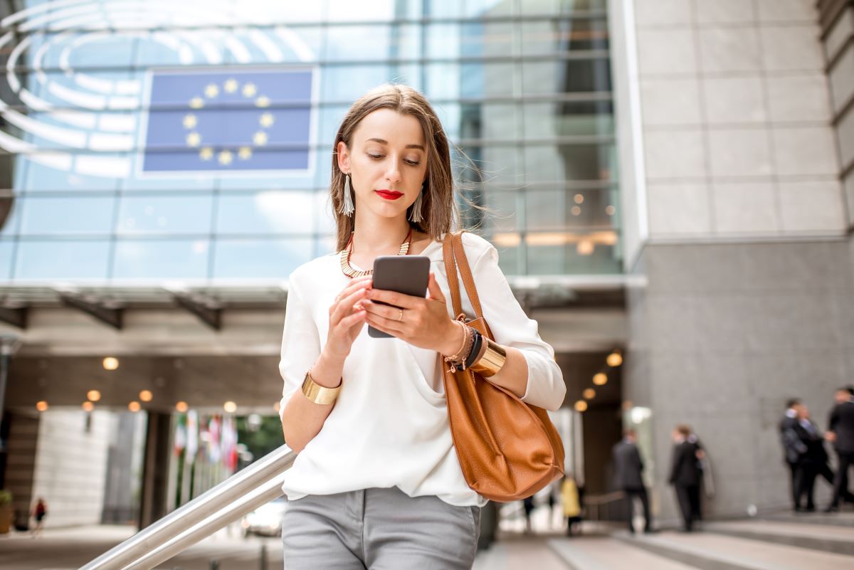 Eine Frau mit langen Haaren, weißer Bluse und grauer Hose, steht vor einem modernen Gebäude mit der Flagge der Europäischen Union, schaut auf ihrem Smartphone auf Neue Calls und trägt eine braune Handtasche. Im Hintergrund sind mehrere Personen zu sehen.
