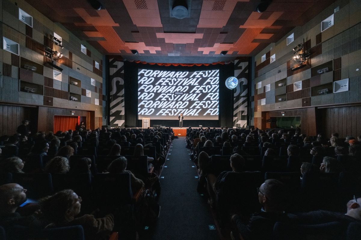 Ein großes Publikum sitzt in einem dunklen Theater mit Blick auf eine Bühne, auf der in fetten weißen Buchstaben FORWARD FESTIVAL steht. Der moderne, holzgetäfelte Raum ist Schauplatz eines interdisziplinären Treffens, das von innovativen Summer School-Sitzungen inspiriert ist.
