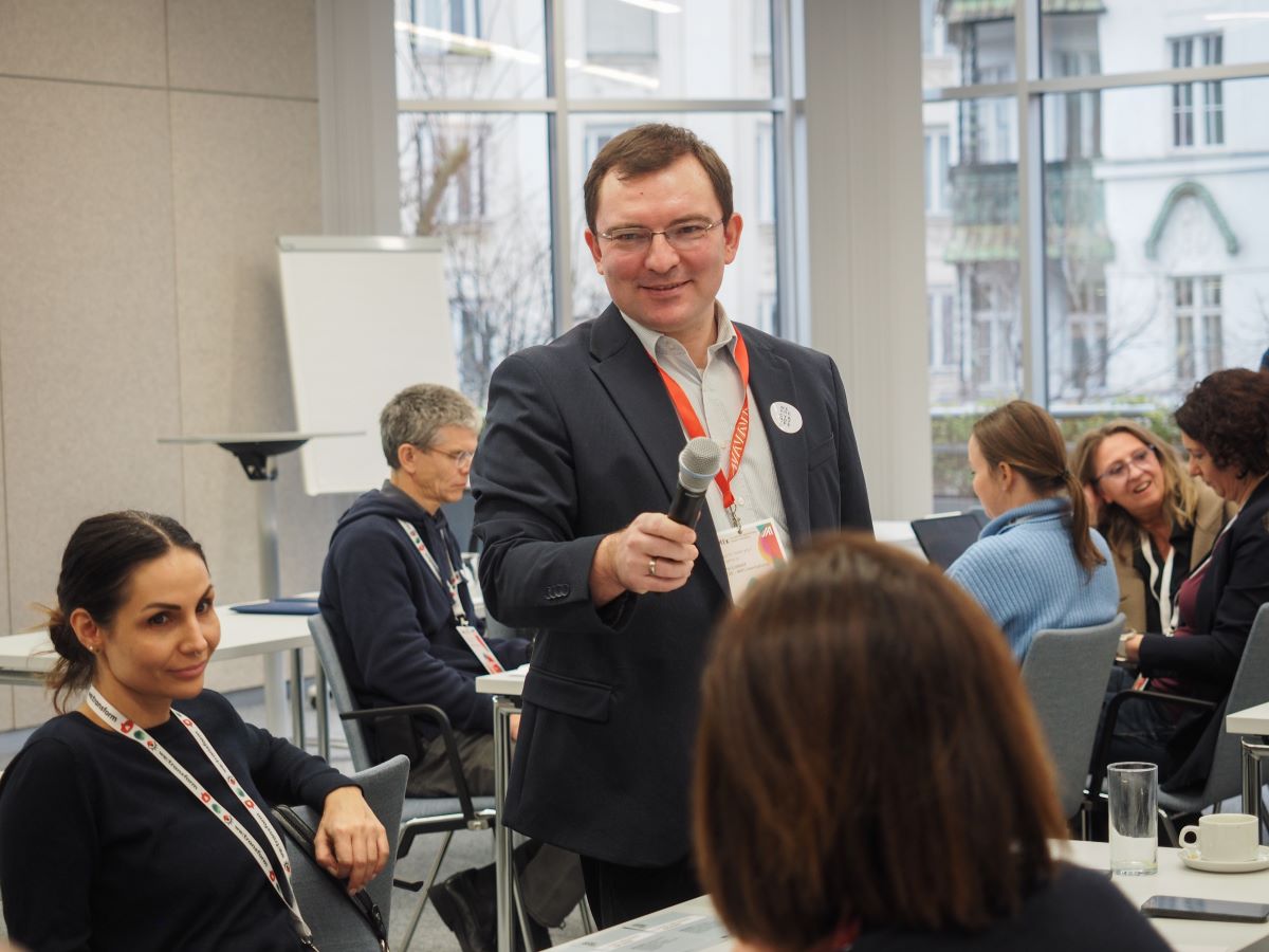 Ein Mann im Anzug, der lächelnd ein Mikrofon in Richtung einer sitzenden Frau hält, unterhält sich mit einer Gruppe in einem hellen, modernen Konferenzraum - eine inspirierende Szene, die den zukunftsorientierten Geist und die Zukunftskompetenzen der heutigen Kreativwirtschaft widerspiegelt.