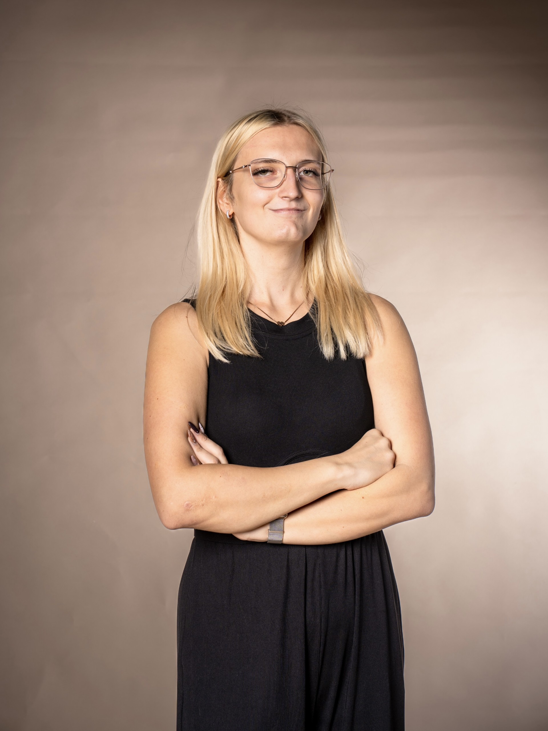 Eine Frau mit langen blonden Haaren und Brille, die ein ärmelloses schwarzes Outfit trägt, steht mit verschränkten Armen und lächelt selbstbewusst und verkörpert die Leichtigkeit von Buchhaltung per App vor einem schlichten beigen Hintergrund.