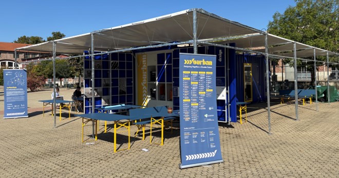 Eine blaue Pop-up-Struktur aus einem Schiffscontainer steht im Freien auf einer gepflasterten Fläche, um die herum gelbe Tische und Bänke stehen. Schilder mit Informationen sind angebracht, und ein paar Leute sind in der Nähe. Im Hintergrund sind Bäume und Gebäude zu sehen.
