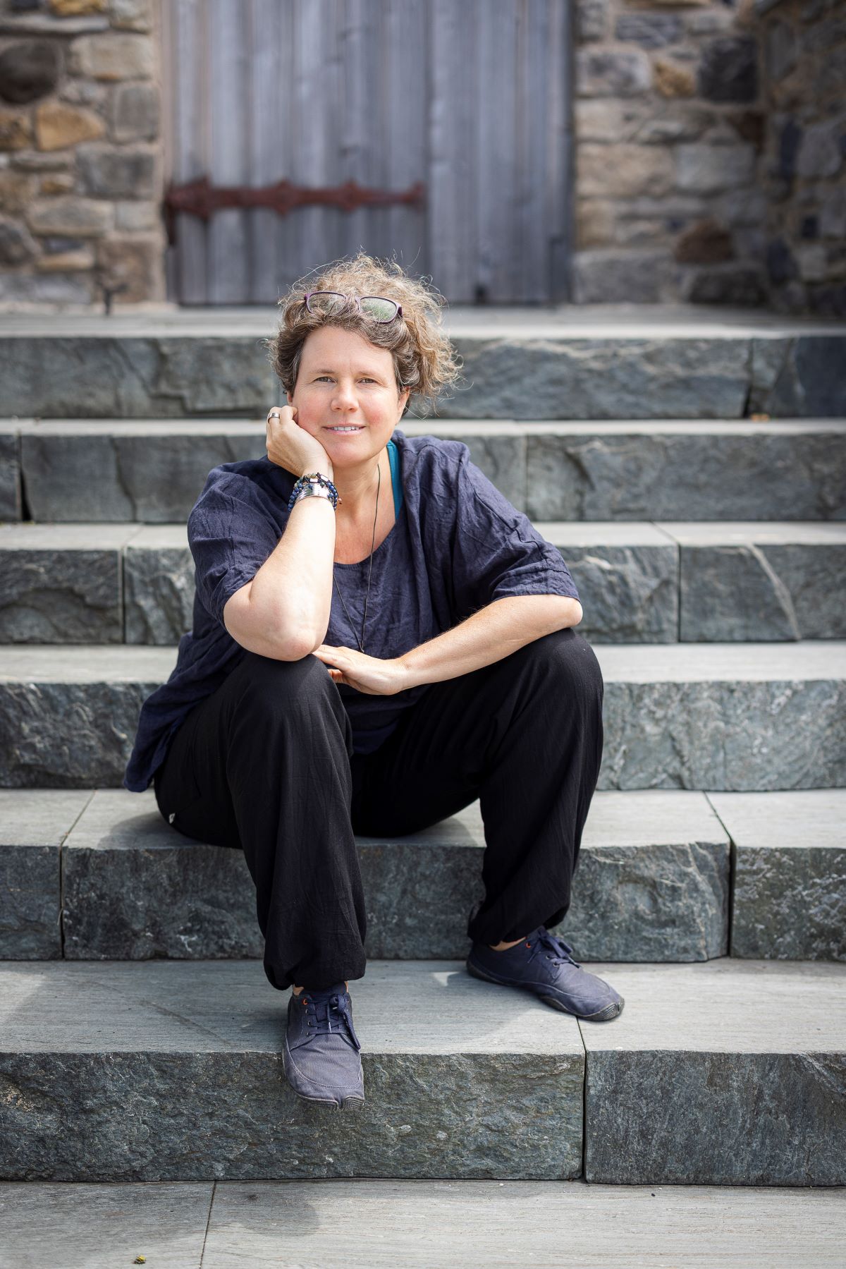 Eine Frau mit kurzen lockigen Haaren, die ein dunkles Hemd und eine dunkle Hose trägt, sitzt lässig auf einer Steinstufe im Freien und stützt ihr Kinn auf ihre Hand. Mit einer Sonnenbrille auf dem Kopf blickt sie in die Kamera - eine entspannte Figur in der Welt der Kreativwirtschaft.