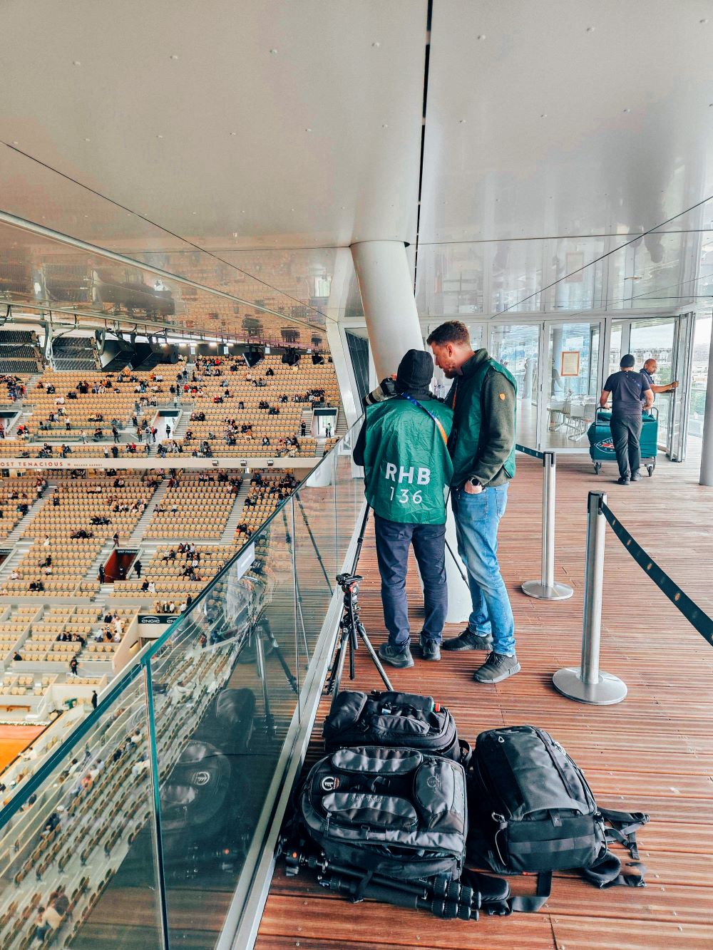 Zwei Personen, möglicherweise Filmemacher wie Peter Jablonowski, stehen auf einem überdachten Balkon mit Blick auf ein Stadion. Kameraausrüstung und Rucksäcke liegen auf dem Holzboden, während sie ihre nächste Zeitraffer-Aufnahme besprechen, während sich die Plätze unter ihnen mit Zuschauern füllen.