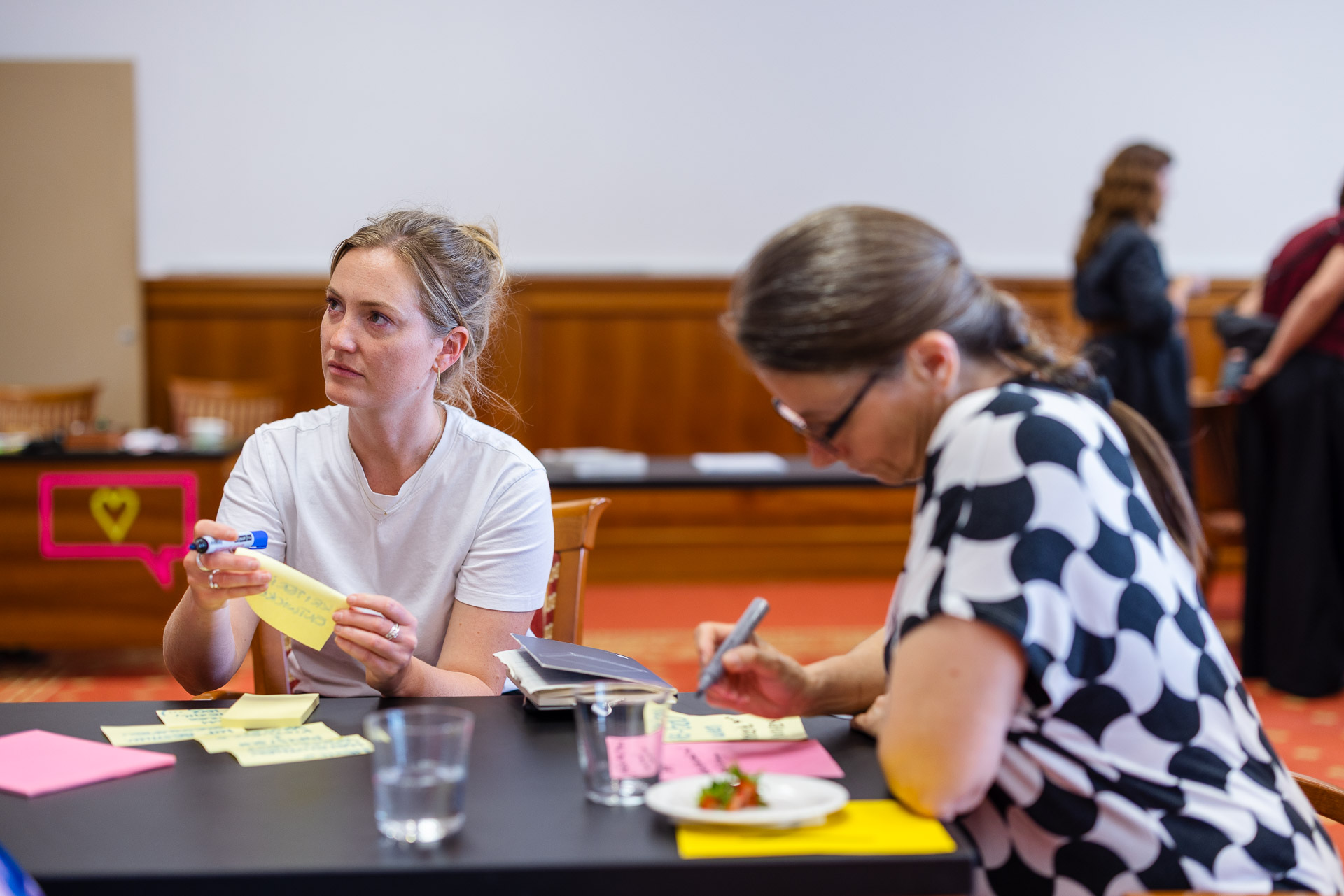 Zwei Frauen sitzen an einem Tisch und schreiben auf Zetteln, eine hält einen Marker und Haftnotizen, die andere schreibt konzentriert. Andere Personen sind unscharf im Hintergrund zu erkennen. Die Umgebung scheint ein Workshop oder eine Gruppensitzung zu sein.