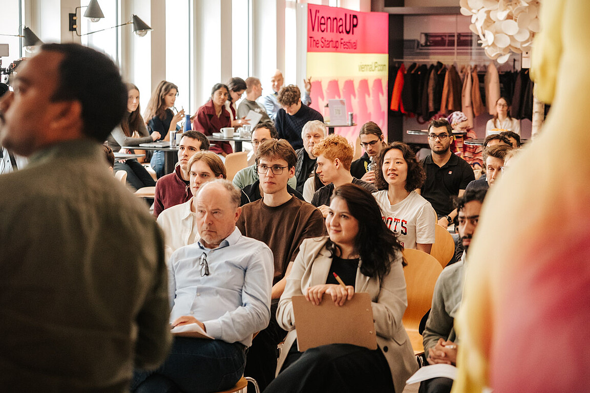 A motley group of people sit in rows and attentively watch a speaker at an indoor event. A sign in the background reads ViennaUP The Startup Festival. The atmosphere is engaging and lively - a true creative catalyst for innovation.