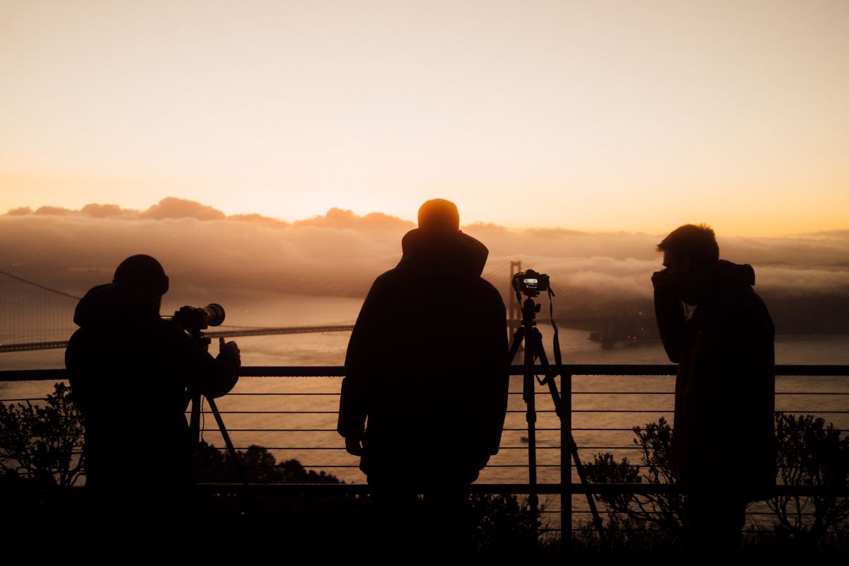 Drei Personen stehen bei Sonnenuntergang an einem Geländer, zwei von ihnen mit Kameras auf Stativen, die einen eindrucksvollen Zeitraffer festhalten, mit Blick auf eine neblige Landschaft mit einer Brücke im Hintergrund. Die Szene ist von hinten beleuchtet, so dass sich die Figuren als Silhouetten abzeichnen.
