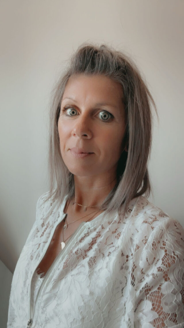 Eine Frau mit glattem, schulterlangem, grauem Haar und grünen Augen steht vor einem schlichten, hellen Hintergrund. Sie trägt eine weiße Spitzenbluse und mehrreihige Ketten, die das ruhige Selbstbewusstsein der KAT-Organisation repräsentieren, während sie in die Kamera schaut.