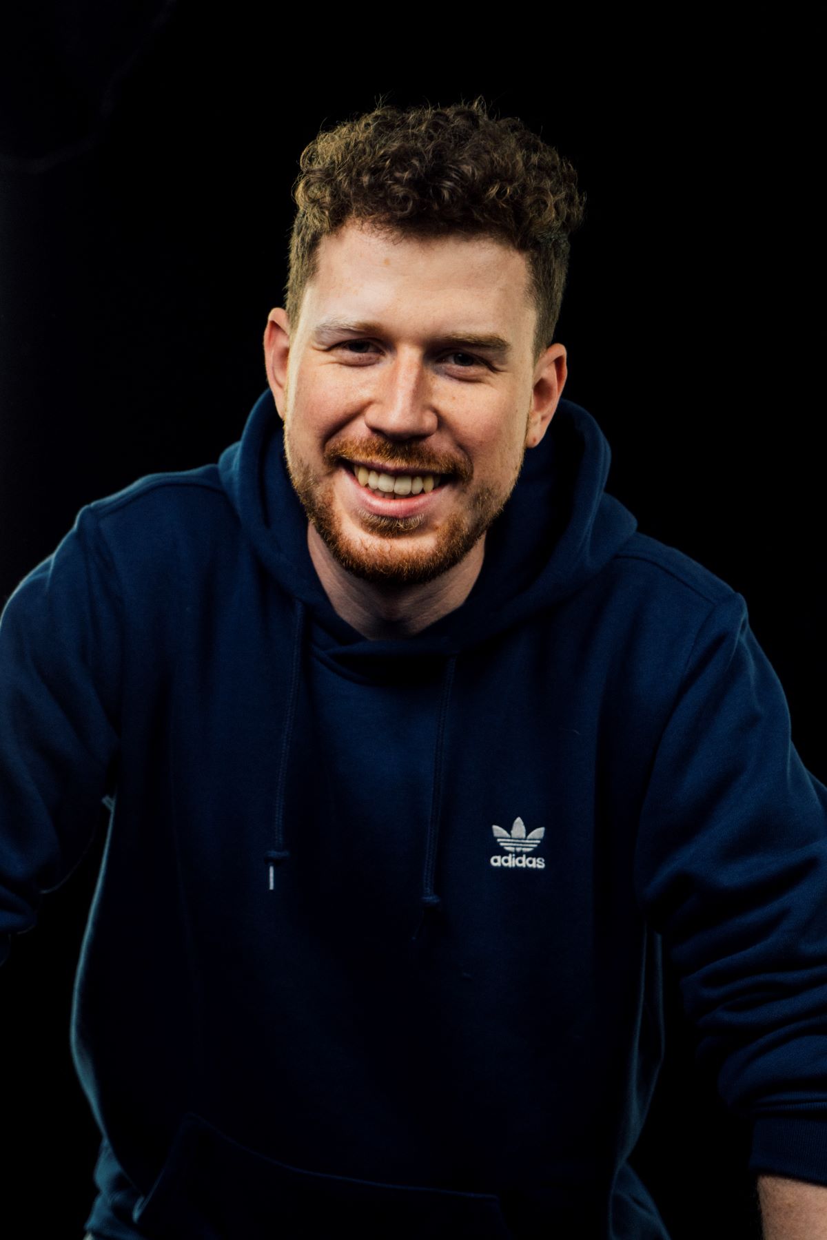 Ein Mann mit kurzen lockigen Haaren und einem Bart l&auml;chelt in die Kamera. Er tr&auml;gt einen marineblauen Adidas-Kapuzenpulli und steht vor einem schwarzen Hintergrund - das k&ouml;nnte Peter Jablonowski sein, der f&uuml;r seine Emmy-Nominierung in Zeitraffer bekannt ist.