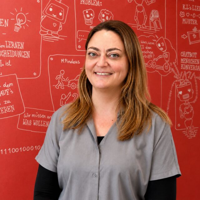 Eine Frau mit langen hellbraunen Haaren, die eine graue Bluse über einem schwarzen Oberteil trägt, steht lächelnd vor einer roten Wand, die mit weißen Cartoon-Zeichnungen und deutschen Texten über Technik und Lernen bei der KAT-Organisation verziert ist.