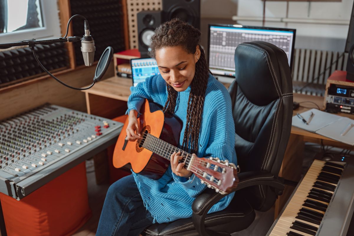 Eine Person in einem blauen Pullover sitzt in einem Musikstudio und spielt auf einer Akustikgitarre. Um sie herum sind Studioausrüstung, ein Mikrofon, Computerbildschirme, auf denen eine Buchhaltungs-App zu sehen ist, eine Tastatur und ein Mischpult zu sehen.