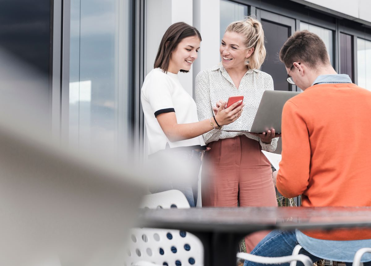 Zwei Frauen stehen draußen, lächeln und schauen auf ein Telefon, während ein Mann in der Nähe sitzt und einen Laptop benutzt. Sie scheinen ein lockeres Gespräch zu führen oder zusammen zu arbeiten.