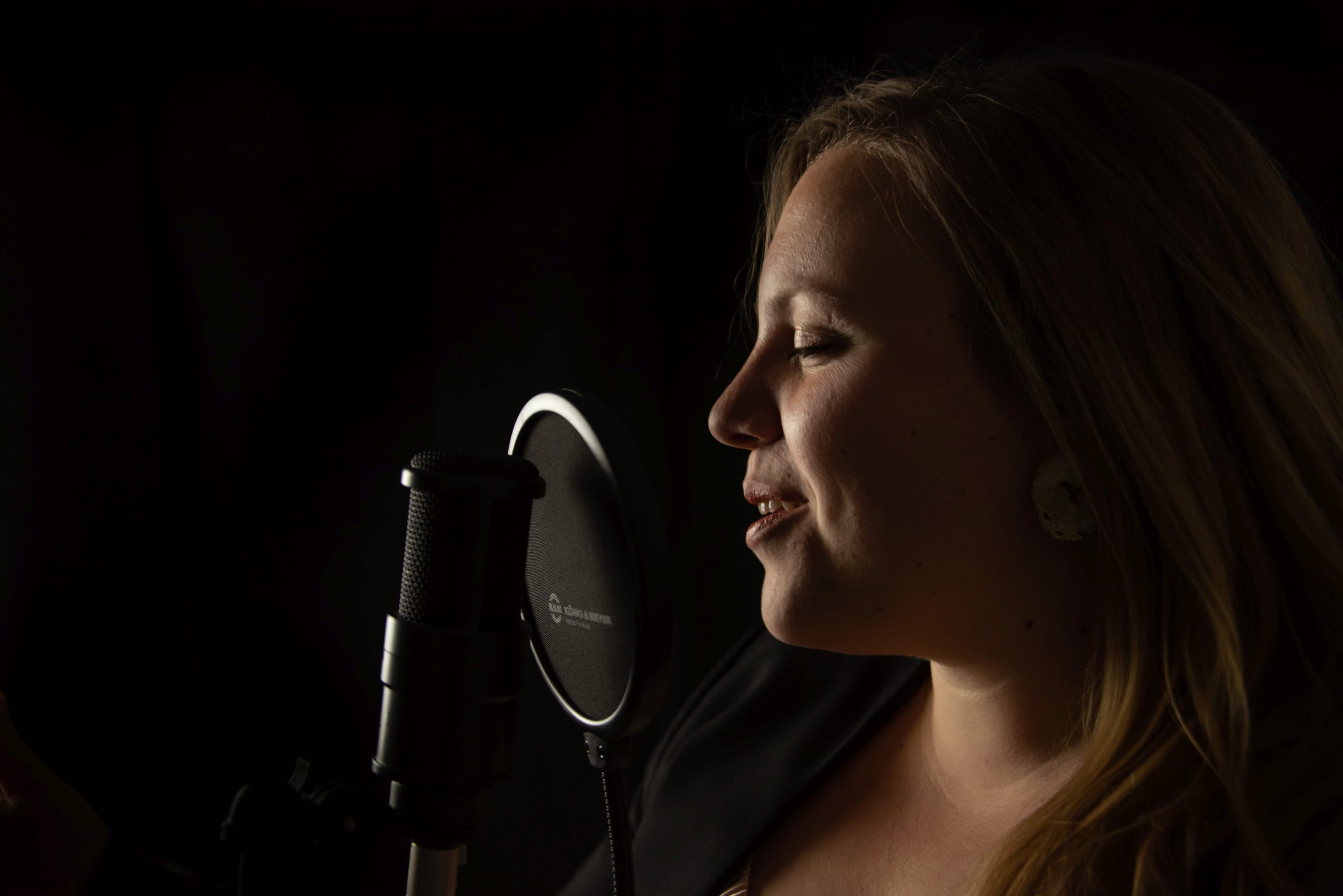 Eine Frau mit langen Haaren, die Melanie Fedl ähnelt, singt in einem schwach beleuchteten Studio in ein Mikrofon mit Pop-Filter. Ihr Gesicht wird vor einem dunklen Hintergrund sanft beleuchtet, während sie nach dem richtigen Ton sucht.