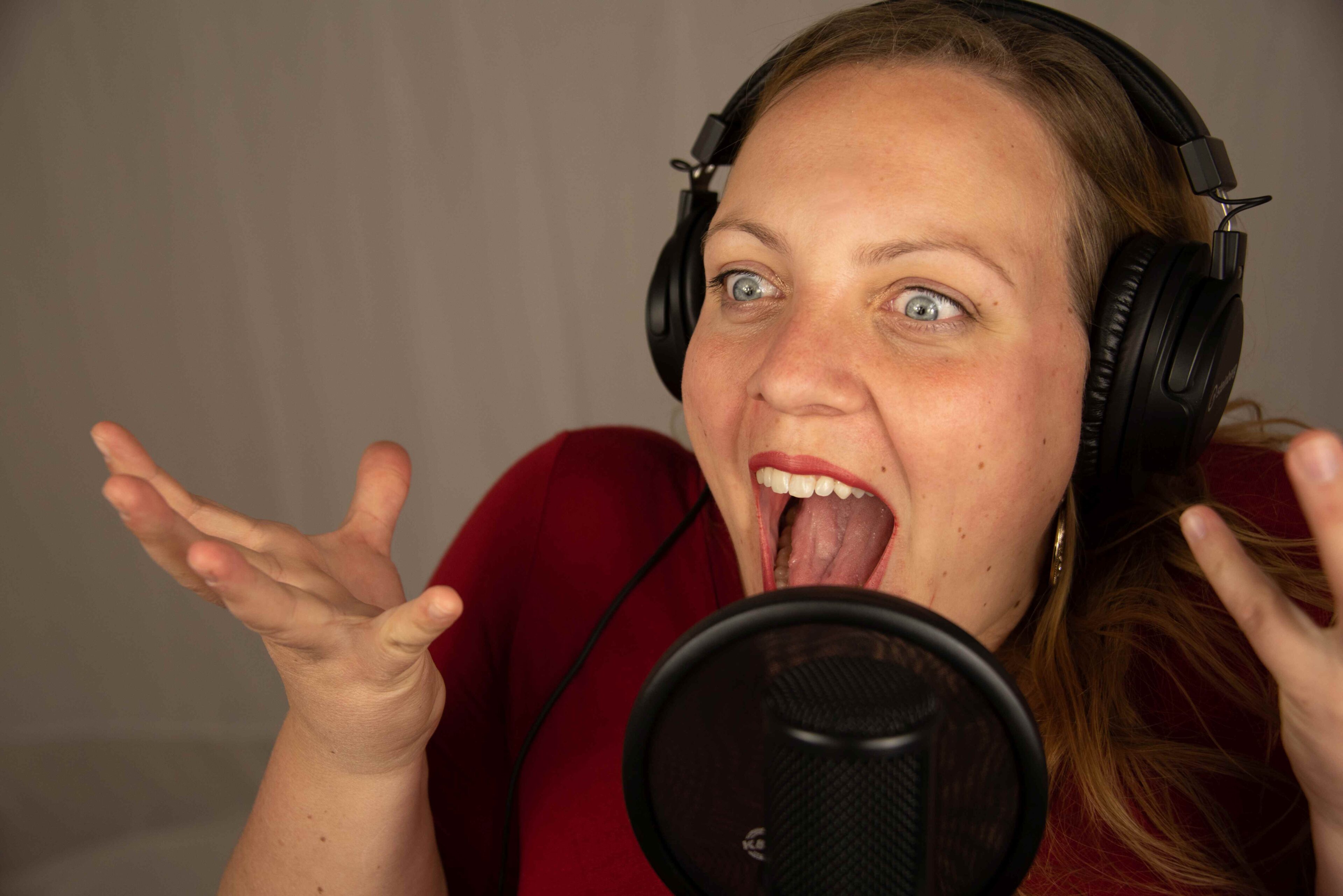 Eine Frau mit Kopfhörern, möglicherweise Melanie Fedl, findet leidenschaftlich den richtigen Ton, während sie mit ausdrucksvoll erhobenen Händen in einem Musik-Aufnahmestudio in ein Mikrofon singt oder spricht.