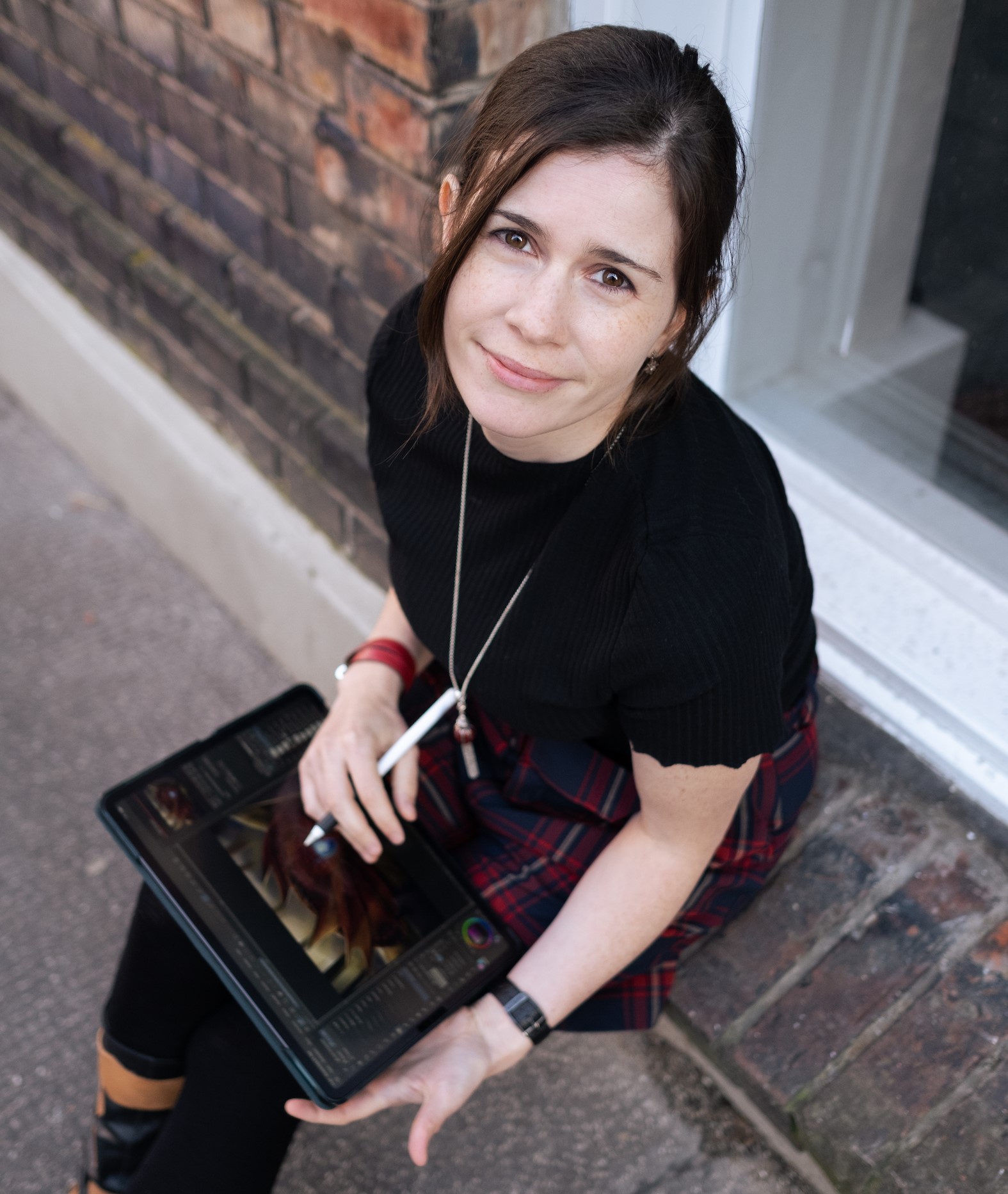 Eine Frau mit braunen Haaren, einem schwarzen Oberteil und einem rotkarierten Rock sitzt vor einem Backsteingebäude und hält ein digitales Tablet und einen Stift in der Hand. Leicht lächelnd blickt sie nach oben, eingetaucht in kreative Welten, während Nadya Knops ihre Gemeinschaft erkundet.