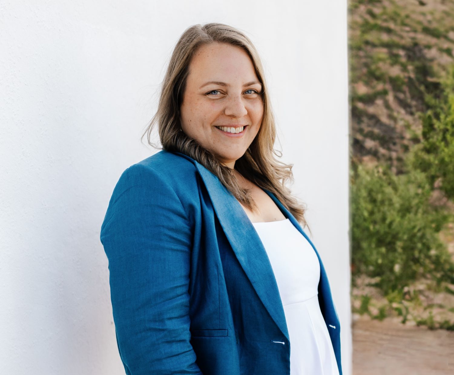 Melanie Fedl, mit langen hellbraunen Haaren, trägt einen blauen Blazer und ein weißes Oberteil. Sie lächelt neben einer weißen Wand, während sie den richtigen Ton inmitten von Grün und sanften Hügeln im Hintergrund einfängt.