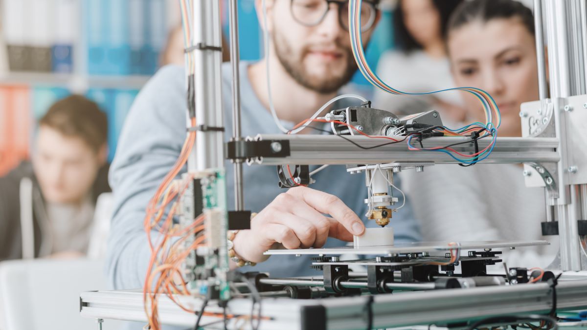 Ein Mann stellt einen 3D-Drucker ein, während eine Frau ihm aufmerksam zusieht. Der Drucker steht im Fokus; man sieht die Kabel und Bauteile, während er Objekte Schicht für Schicht herstellt, während im Hintergrund andere Menschen in einer hellen, modernen Umgebung arbeiten.