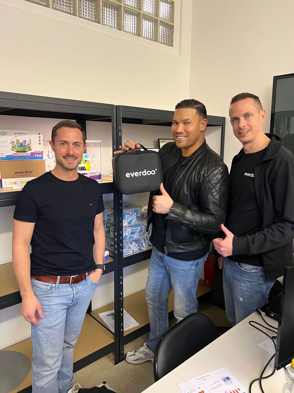 Drei Männer stehen in einem Büro neben Regalen mit Produkten. Der Mann in der Mitte, Patrick Weissinger, hält eine schwarze Everdoo-Tasche in der Hand und streckt den Daumen nach oben. Alle lächeln und sind leger in Jeans und schwarzen Hemden oder Jacken gekleidet; sie strahlen Kreativität aus, während sie praktische Lösungen jenseits der Wegwerfmentalität präsentieren.