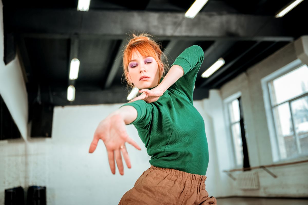 Eine Frau mit roten Haaren, die ein grünes Oberteil und eine braune Hose trägt, tanzt anmutig in einem Studio mit großen Fenstern und schwarzen Deckenbalken. Ihre Augen sind geschlossen, und ein Arm ist nach vorne ausgestreckt – damit verkörpert sie den Geist der Kreativität, der auf dem European Creative Industries Summit oft gewürdigt wird.