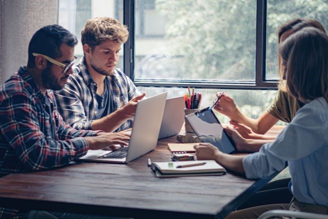 Vier Personen sitzen mit Laptops und Tablets an einem Holztisch und arbeiten gemeinsam an einem Projekt in der Nähe großer Fenster, durch die Tageslicht hereinfällt.