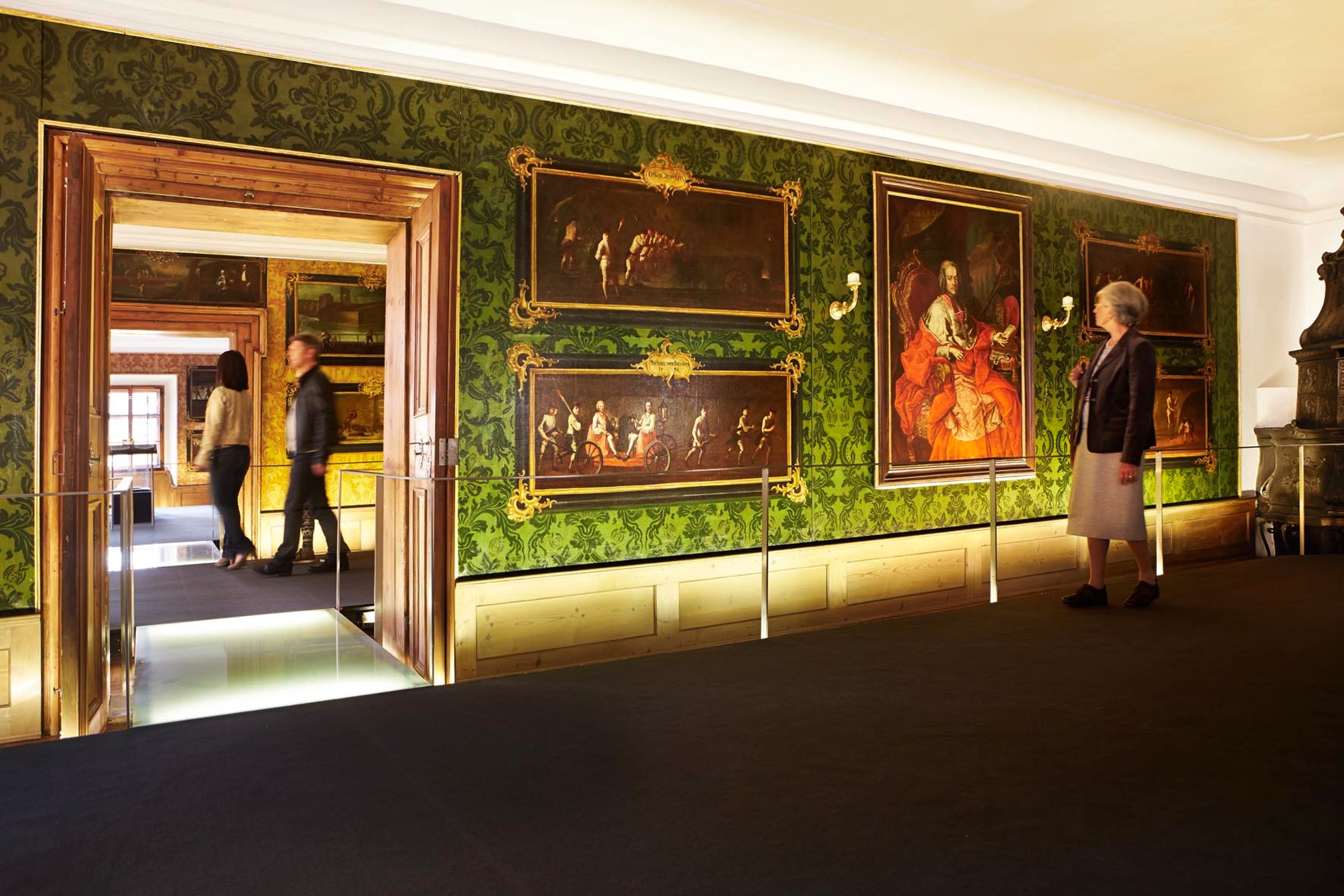 Eine Frau betrachtet klassische Gemälde – vielleicht betrachtet sie jedes einzelne als einen kulturellen Messwert –, die an einer grün gemusterten Wand in einem hell erleuchteten Museumsraum hängen, während im Hintergrund zwei Personen durch eine Holztür gehen.