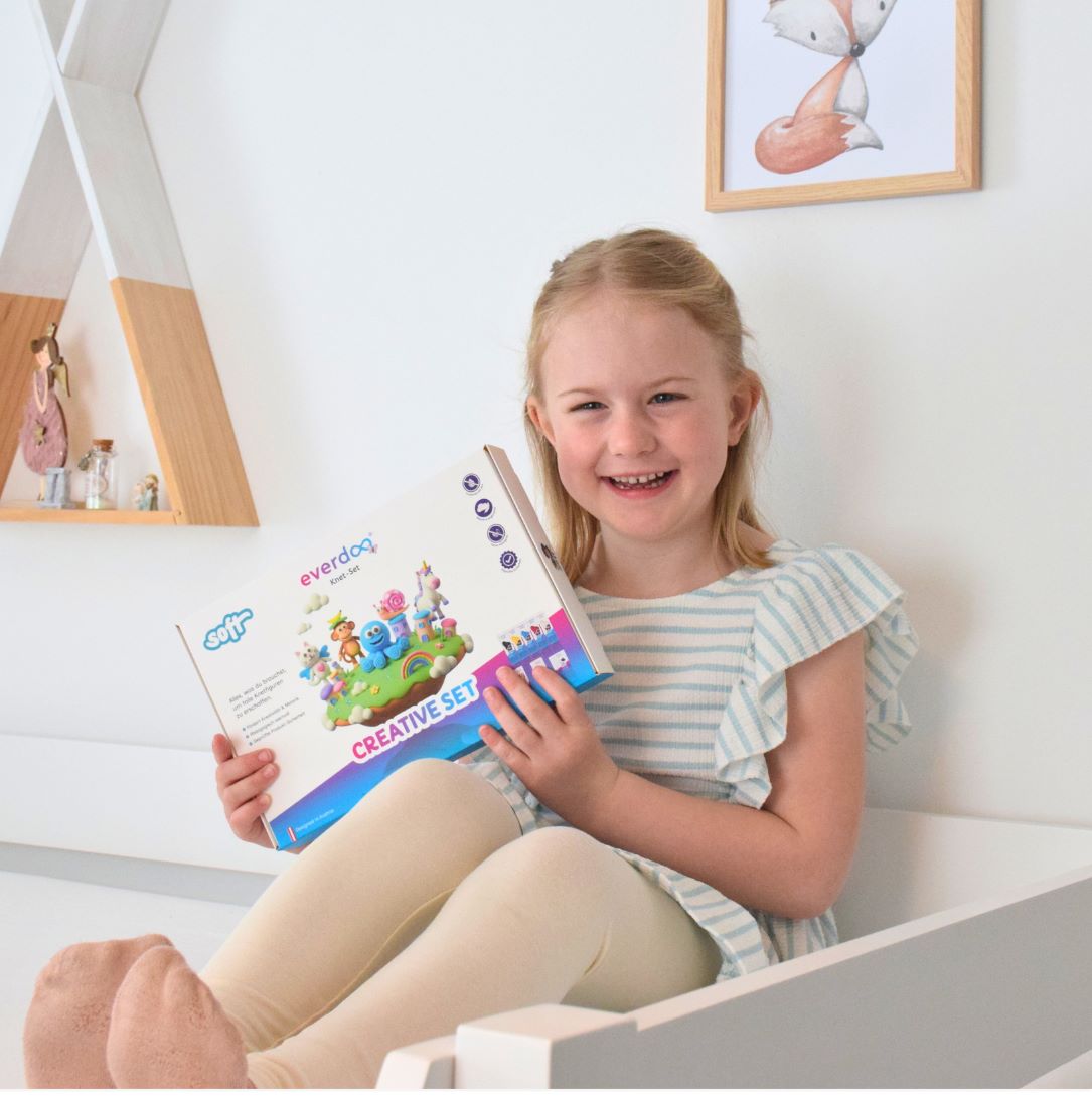 Ein lächelndes Mädchen in einem gestreiften Shirt sitzt auf einem Bett und hält eine bunte Bastelbox in der Hand – bereit, ihrer Kreativität freien Lauf zu lassen. Regale und eine gerahmte Tierillustration verleihen dem Hintergrund zusätzlichen Charme und lassen eine Umgebung erahnen, die die Fantasie anregt und sich von der Wegwerfmentalität abwendet, ganz im Sinne von Patrick Weissinger.