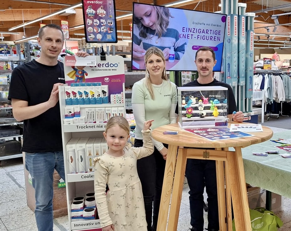 Drei Erwachsene und ein kleines Mädchen stehen in einem Geschäft neben einem Ausstellungsstand, auf dem Mal- und Bastelbedarf präsentiert wird. Voller Neugier und Kreativität zeigt das Mädchen auf Modelliermasse, während im Hintergrund ein Everdoo-Werbeplakat und ein Fernseher zu sehen sind.