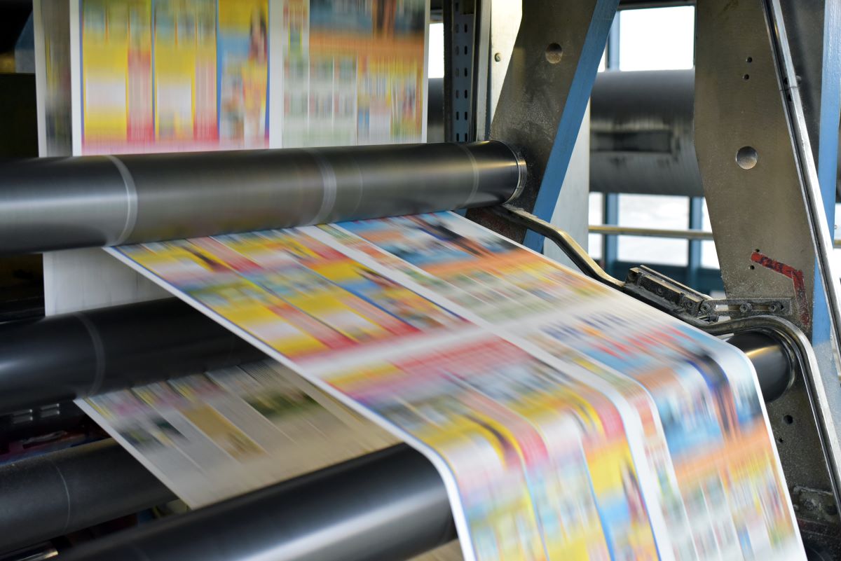 Eine Nahaufnahme einer Druckmaschine von Druck in Betrieb, bei der bunte Papierbögen rasant durch die Walzen laufen und die Präzision und Geschwindigkeit der modernen Medienproduktion verdeutlichen.