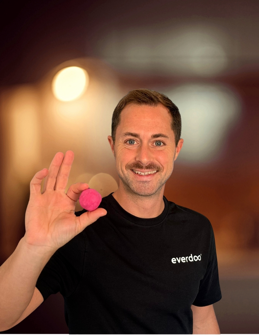 Ein lächelnder Mann in einem schwarzen Everdoo-Hemd, der vielleicht die Kreativität von Patrick Weissinger verkörpert, hält einen kleinen rosa Ball zwischen den Fingern, vor einem sanft unscharfen braunen Hintergrund mit hellen Lichtern.