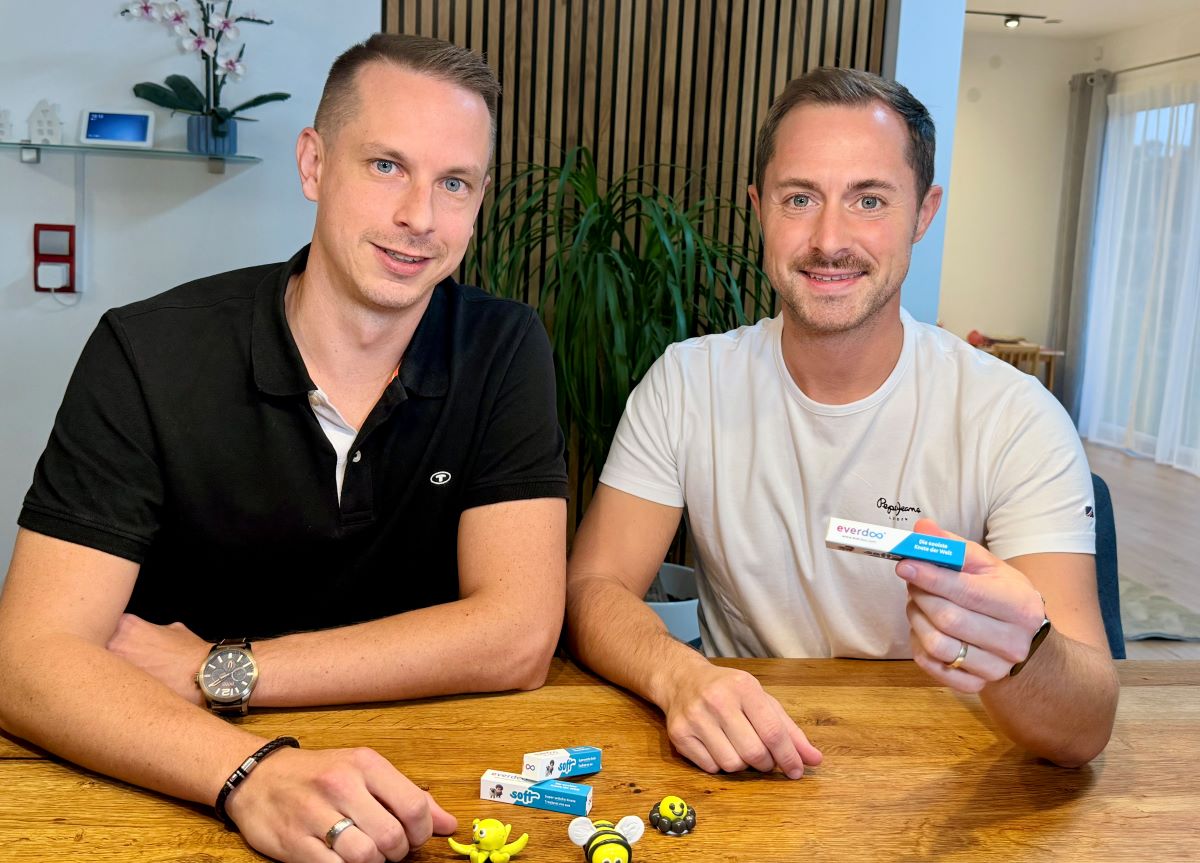 Zwei Männer sitzen an einem Holztisch, auf dem kleines Spielzeug und Verpackungen von Medizinprodukten liegen, und beweisen damit ihre Kreativität. Der eine Mann trägt ein schwarzes Hemd, der andere ein weißes – es handelt sich um Patrick Weissinger –, der in einem hellen, modernen Raum lächelt und eine Schachtel mit der Aufschrift „medeasy“ in der Hand hält.
