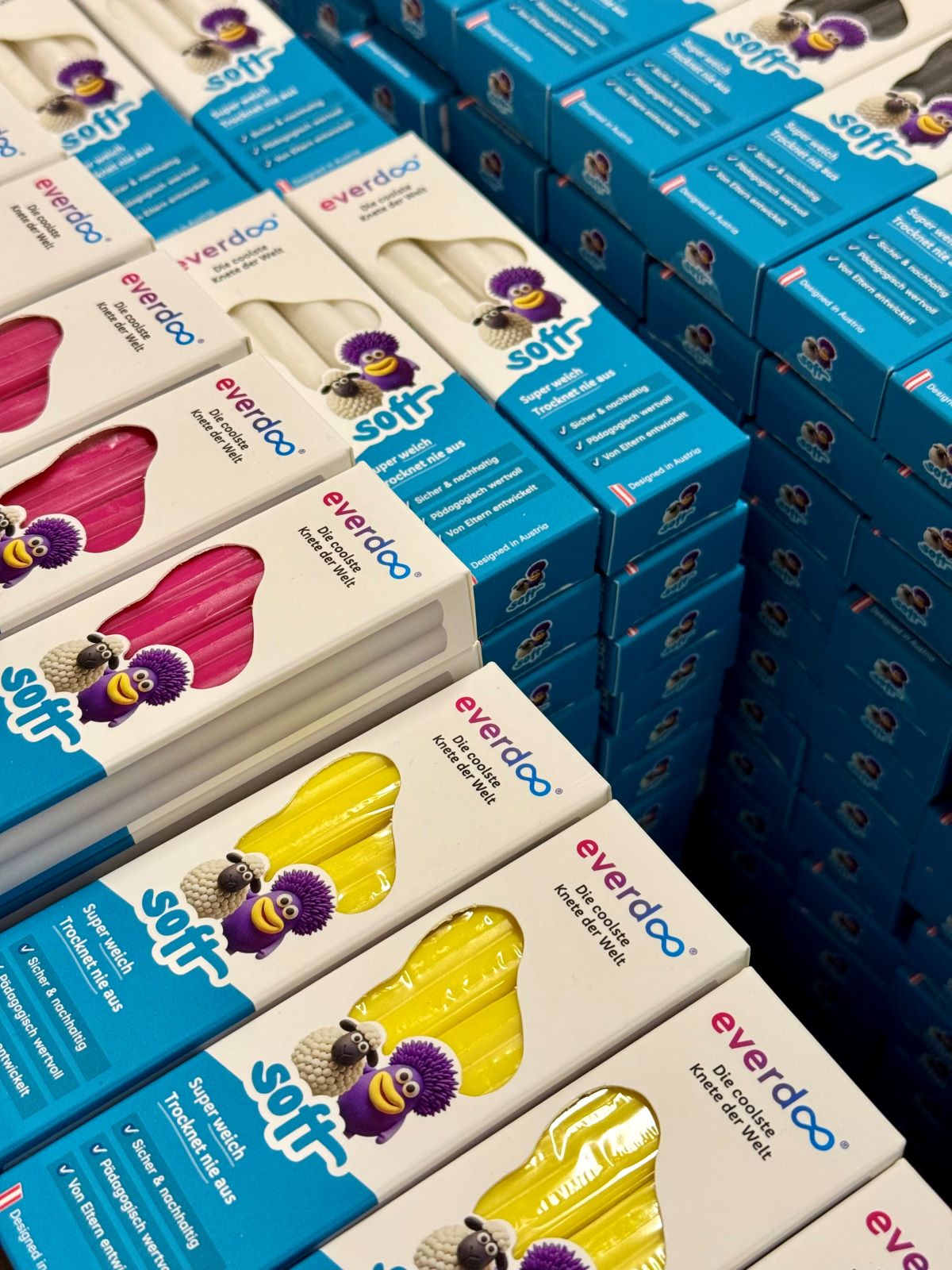 Stapel von Kartons der Marke „everdoo“ in Blau, Rosa und Gelb, auf denen jeweils die Abbildung einer Zeichentrickfigur und das Wort „soft“ zu sehen sind. Die Kartons, die in einem Ladenregal stehen und offenbar Schwämme oder Reinigungsprodukte enthalten, sorgen für einen Hauch von Kreativität und thematisieren zugleich moderne Bedenken hinsichtlich der Wegwerfmentalität.