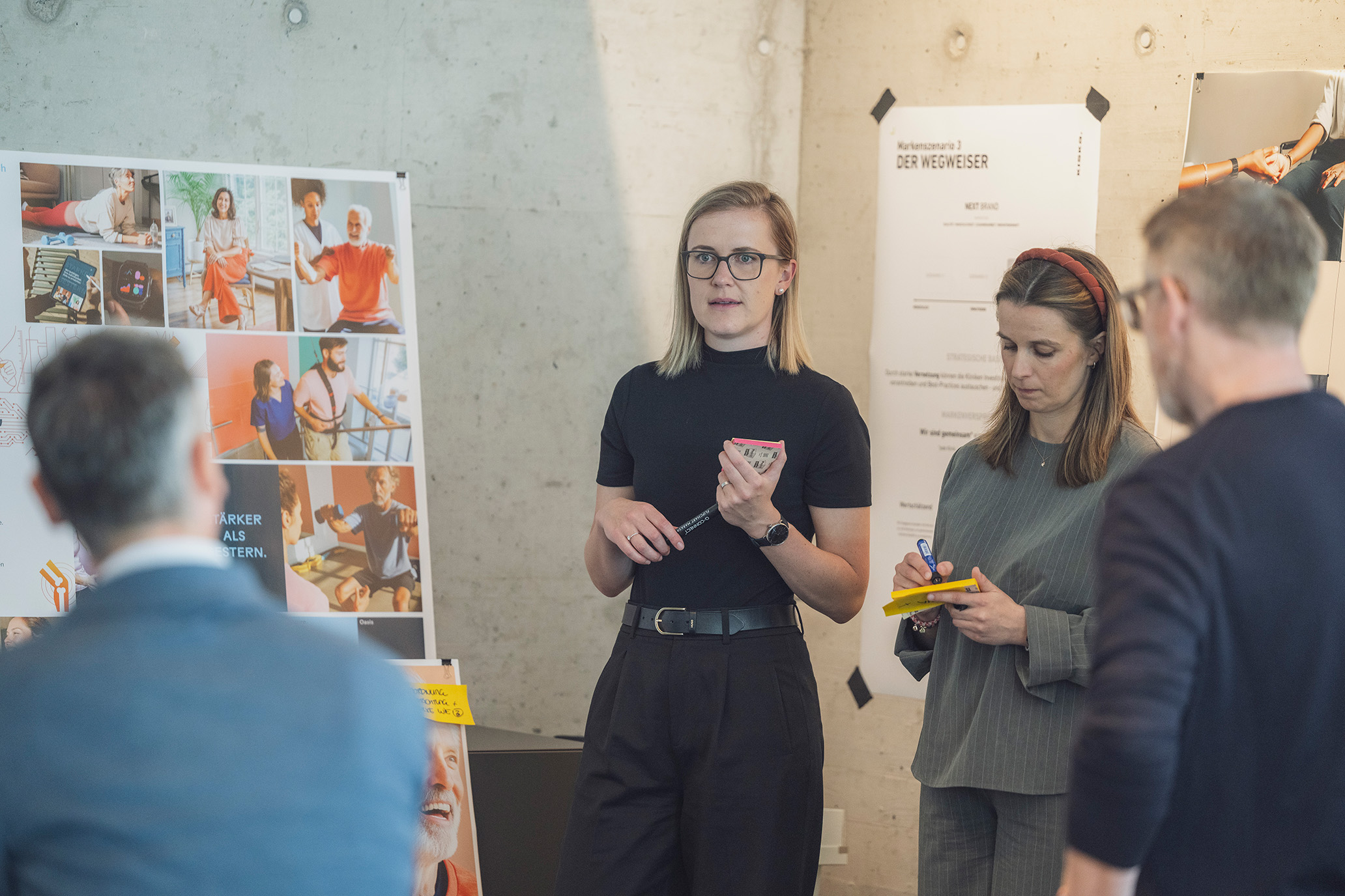 In einem modernen, auf Zusammenarbeit ausgerichteten Büro stehen zwei Frauen und sprechen mit zwei sitzenden Personen; eine von ihnen hält neben einer Tafel mit Fotos eine Präsentation. Post-it-Zettel und Dokumente sind zu sehen, was die Atmosphäre des Unternehmenswandels widerspiegelt, während das Team Strategien zur Gestaltung einer neuen Identität erörtert.