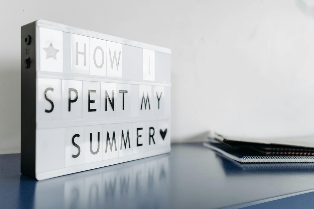 Auf einem Leuchtkasten steht „HOW I SPENT MY SUMMER“ zusammen mit einem Herzsymbol, was auf neue Perspektiven hindeutet, die während der Ferien gewonnen wurden. Daneben liegen ein paar gestapelte Notizbücher auf einer blauen Fläche vor einer schlichten weißen Wand.
