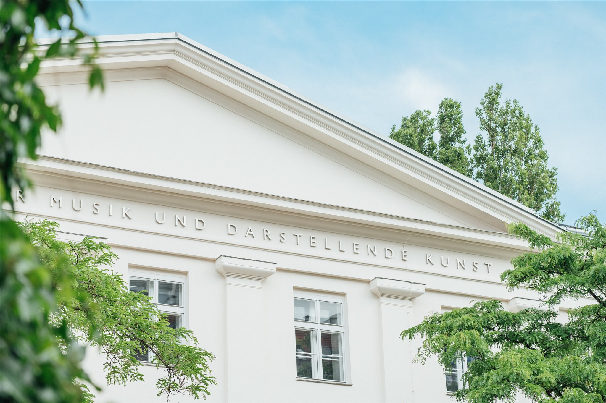 Ein weißes, neoklassizistisches Gebäude, das teilweise von grünen Bäumen verdeckt wird; nahe der Dachlinie ist die Aufschrift „MUSIK UND DARSTELLENDE KUNST“ zu sehen. Der Himmel darüber ist klar und blau, was ein Gefühl kreativer Kraft vermittelt und auf ein Umfeld hindeutet, das von ökosozialem Wandel geprägt ist.