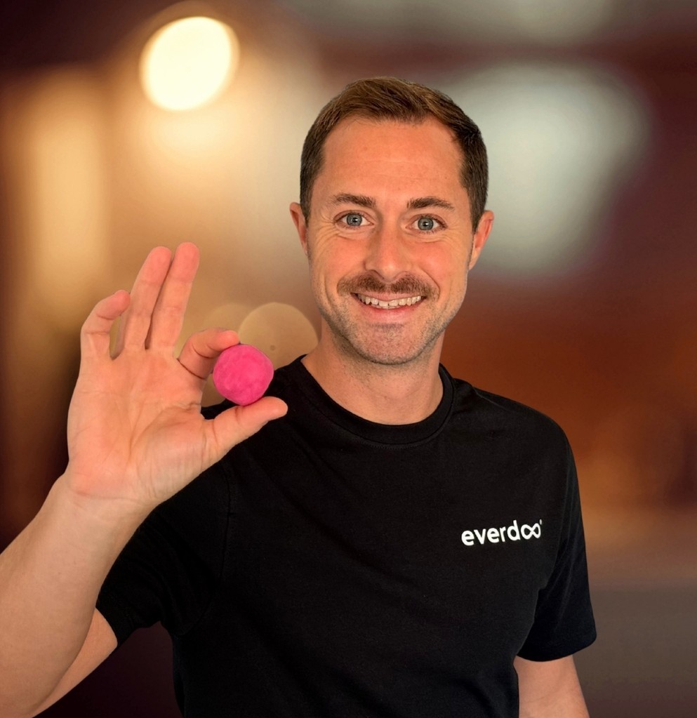 Ein lächelnder Mann, Patrick Weissinger, der ein schwarzes Everdo-Hemd trägt, hält einen kleinen rosa Ball zwischen den Fingern. Der Hintergrund ist sanft unscharf und in warmes Licht getaucht, was eine Atmosphäre der Kreativität und der nachdenklichen Absicht vermittelt.