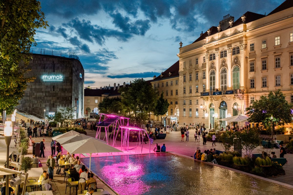 Ein belebter Stadtplatz in der Abenddämmerung, auf dem Menschen spazieren gehen und am Rand eines Wasserbeckens sitzen, beleuchtete Schaukeln mit rosa Lichtern sowie historische und moderne Gebäude im Hintergrund, darunter das mumok – ein Bild, das die pulsierende Atmosphäre des MuseumsQuartiers Wien einfängt, das sich auf Veranstaltungen wie die „Avant Première 2027“ freut.