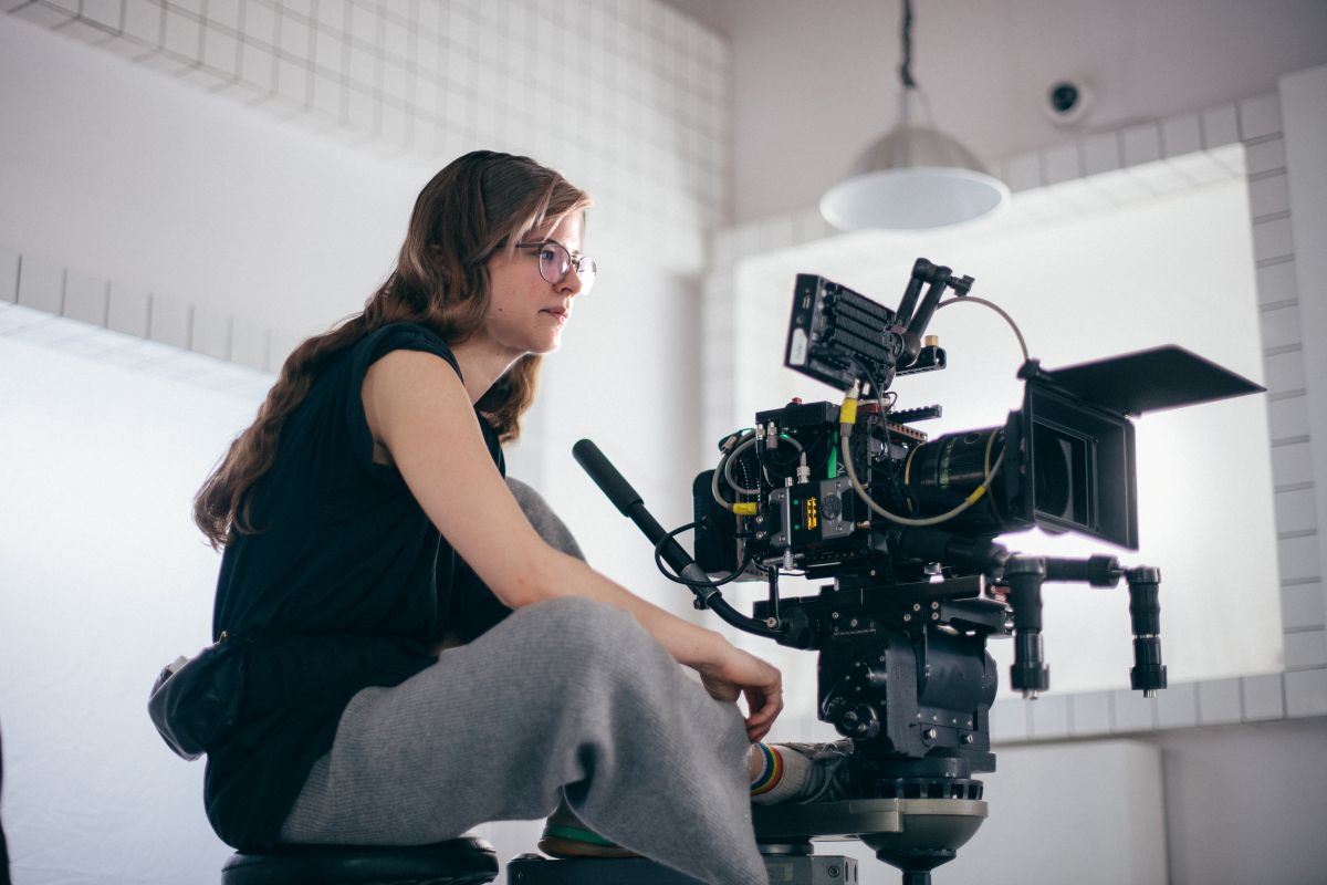 Eine Person mit langen Haaren und Brille bedient eine professionelle Videokamera in einem hellen, modernen Innenraum mit weißen Fliesenwänden und einer Hängelampe und fängt dabei die lebendige Atmosphäre der zeitgenössischen Filmkultur ein – vielleicht dokumentiert sie Projekte, die durch das Elisabeth-Büttner-Stipendium gefördert werden.