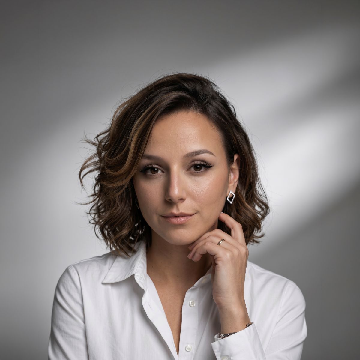 A woman with short, wavy brown hair, wearing a white button-down shirt, poses against a gray background, touching her face with one hand and looking confidently into the camera.