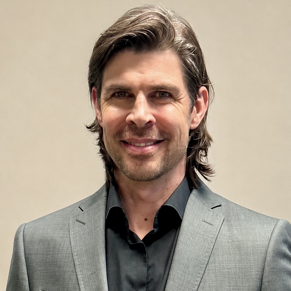 A man with medium-length brown hair, wearing a gray blazer over a black shirt, smiles at the camera against a plain, light-colored background.