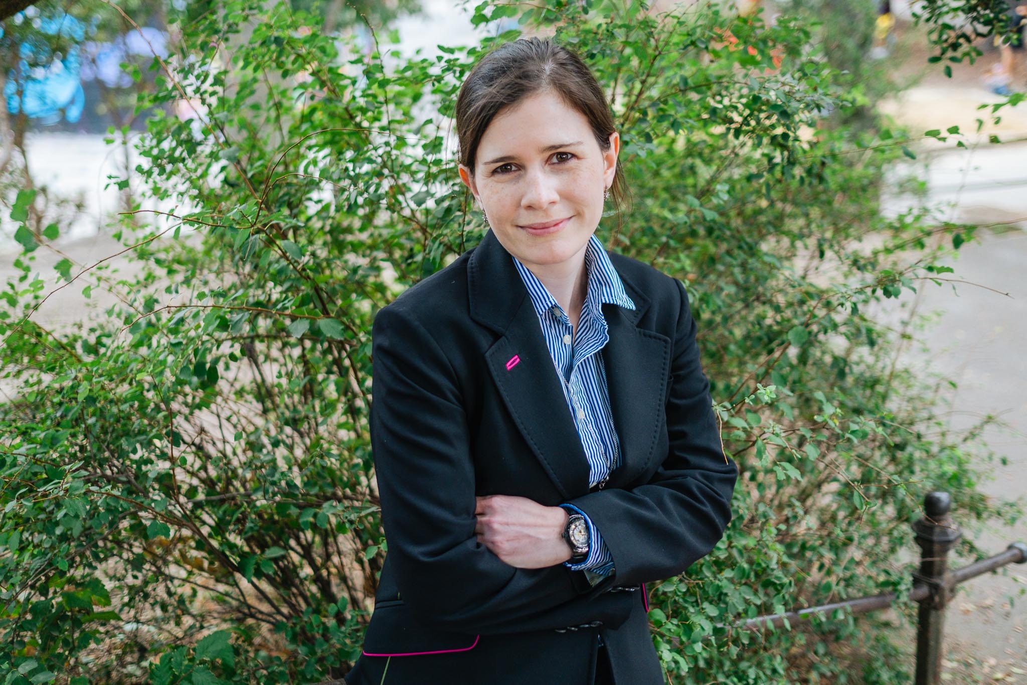 A woman wearing a black blazer and a blue-striped shirt is standing outdoors, her arms crossed, smiling slightly. Green bushes and trees can be seen in the background.