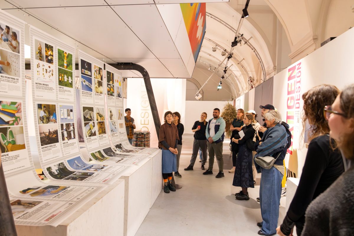 Eine Gruppe von Besuchern betrachtet eine umfangreiche Ausstellung mit gedruckten Plakaten und Bildern in einem modernen, hell beleuchteten Galerieraum mit hohen Gewölbedecken, in dem die Ausstellung Themen wie „Next Gen Design“ und „Futures Worth Living“ in den Mittelpunkt stellt.