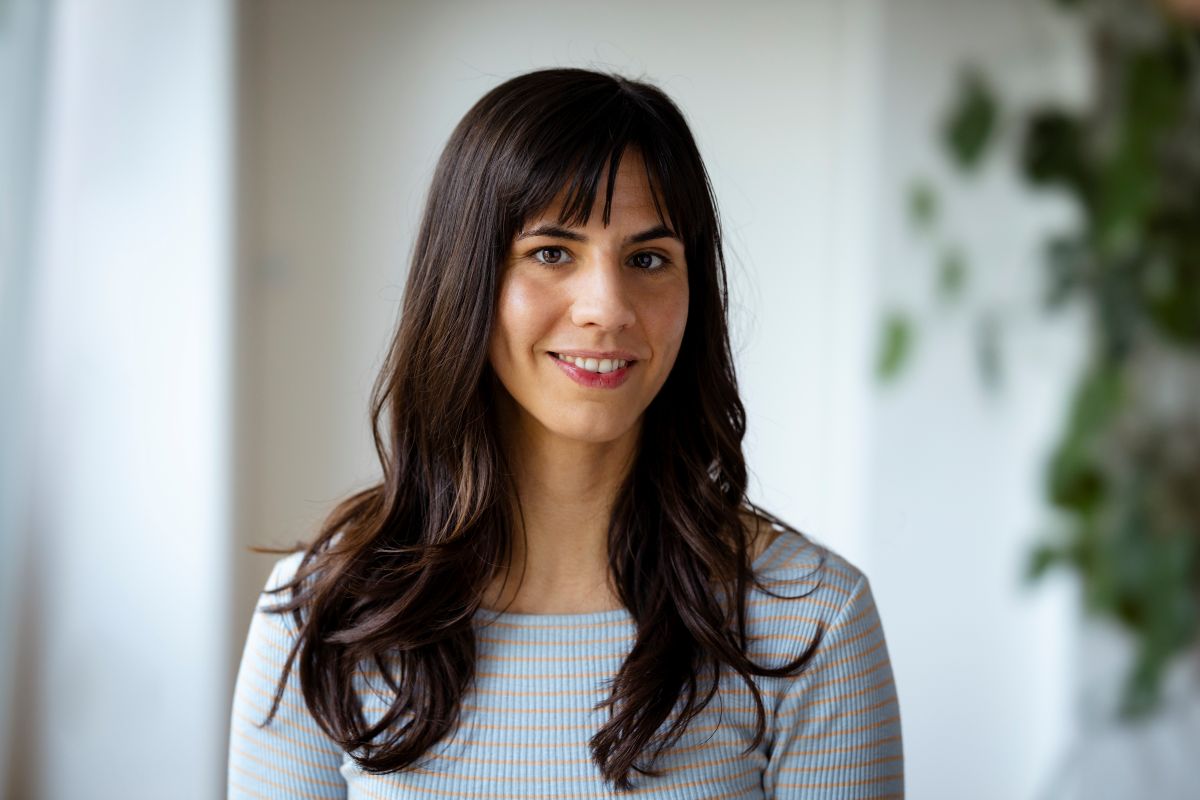 A woman with long brown hair and bangs smiles gently while wearing a light-colored, striped top. She is standing indoors in front of a blurred background featuring green plants, which conveys the warmth of community and the inspiration of creative worlds.